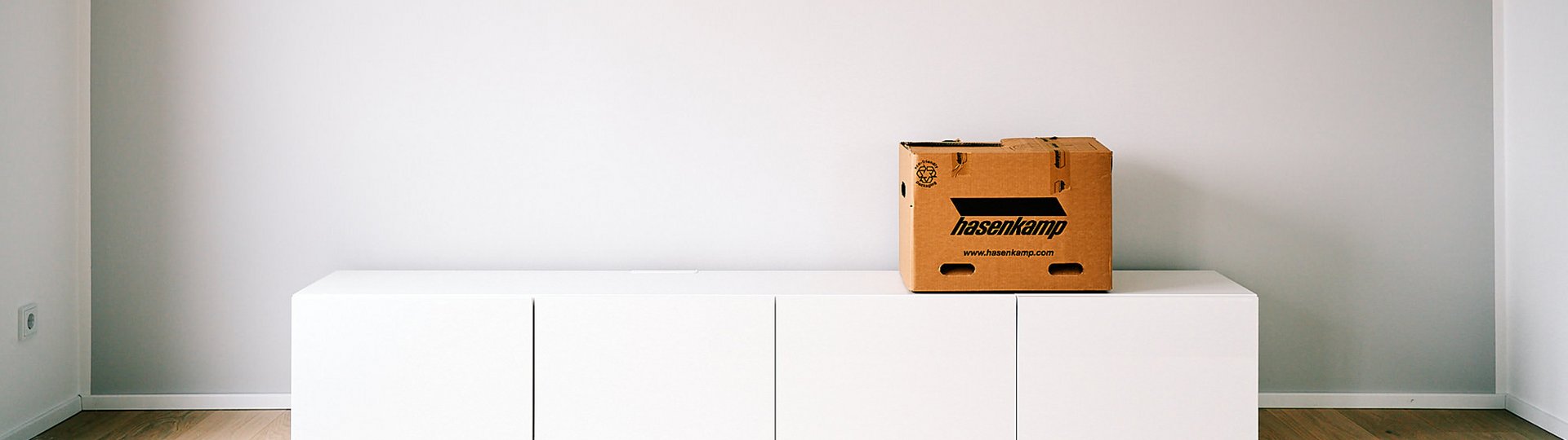 Ein leerer Raum mit einem Sideboard, auf dem eine Umzugskiste mit hasenkamp Aufdruck steht.
